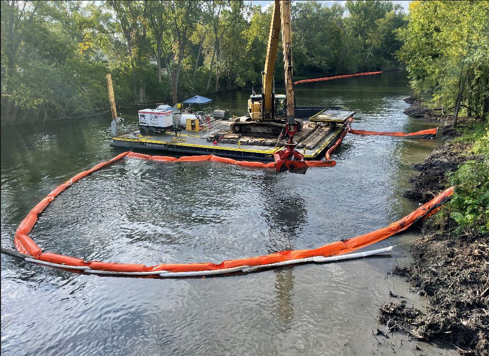 An excavator mounted on a barge dredges river sediment inside a long orange containment boom, green trees on both banks.